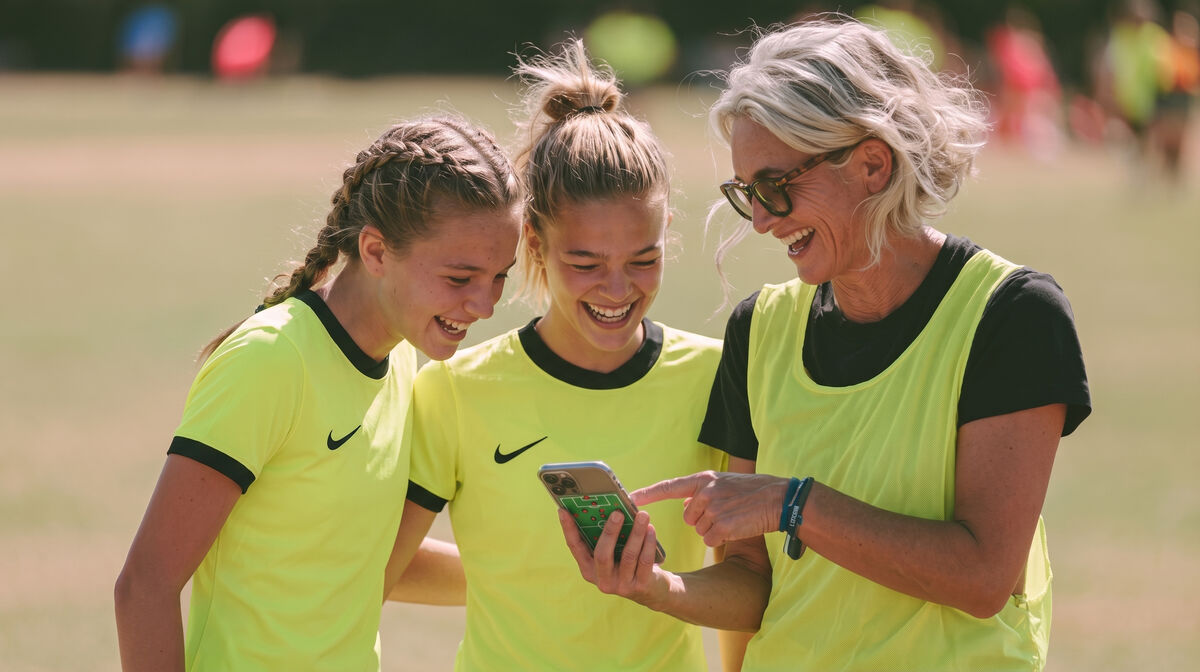 A coach and two young soccer players smiling while looking at stats on a phone during practice